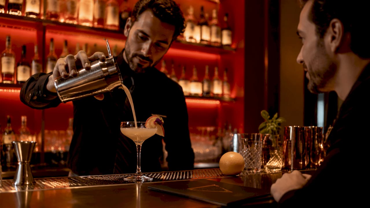 In een gezellige bar staat de barman een kleurrijke cocktail in te schenken.