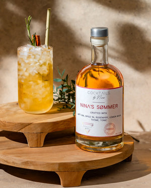 Nina's Sømmers cocktail bottle with a glass of drink, garnished with herbs and spices on wooden coasters.-Cocktails by Nina