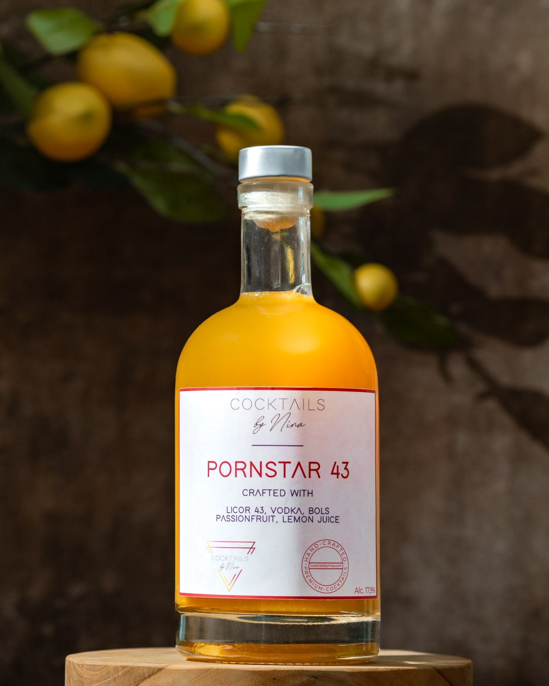 Pornstar 43 cocktail in a clear bottle with a yellow liquid, featuring a white label and wooden base, surrounded by lemons.-Cocktails by Nina